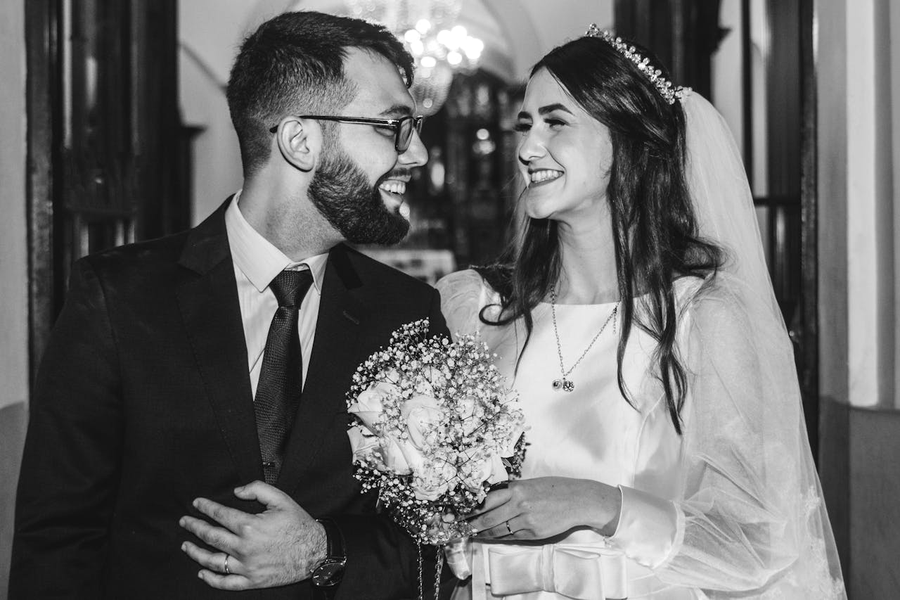 A joyful bride and groom smile in an elegant black and white wedding portrait indoors.